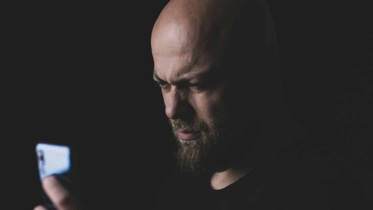 A bald man with a beard stares intensely at a glowing smartphone in a dark room.