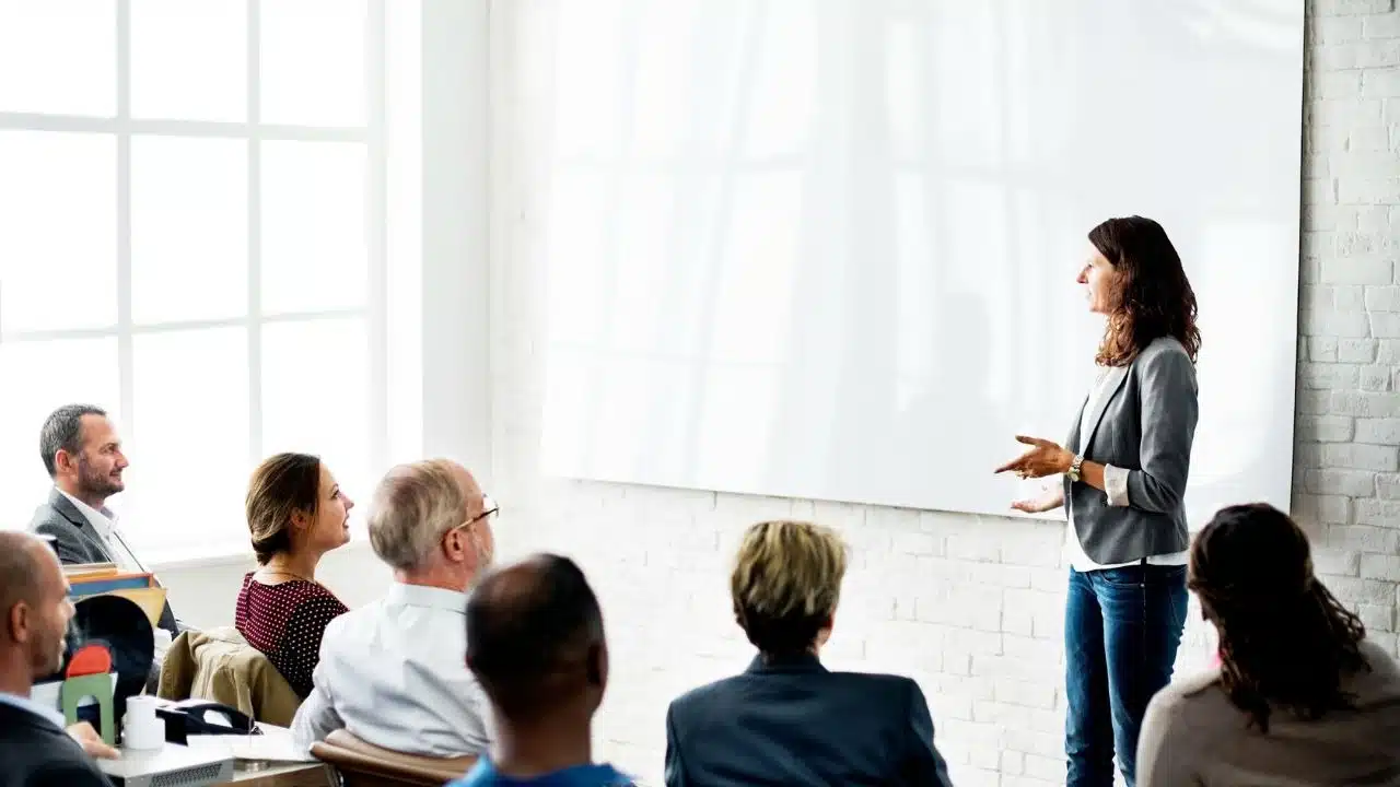 A woman stands and gestures while speaking to a seated audience in front of a whiteboard.