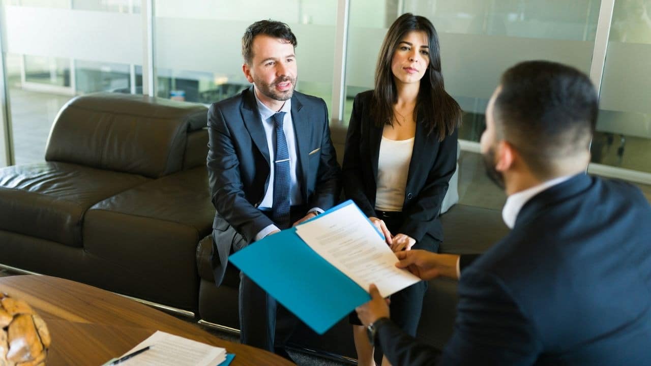 A man and woman in suits sit on a sofa looking at a legal document.
