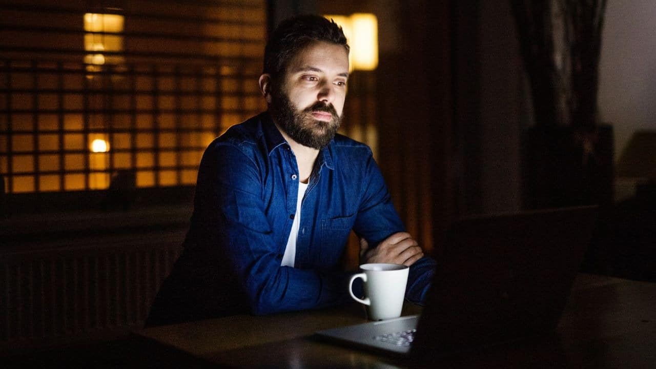 A bearded man sits at a laptop in a dark room with a mug nearby.
