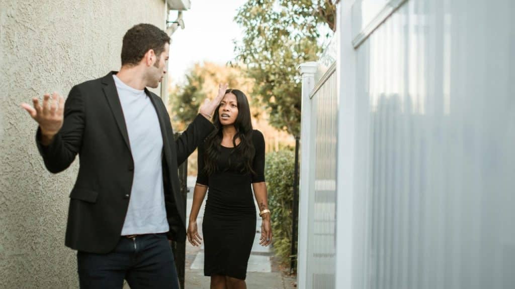 A man in a blazer gestures with his hands while a woman in black looks on.