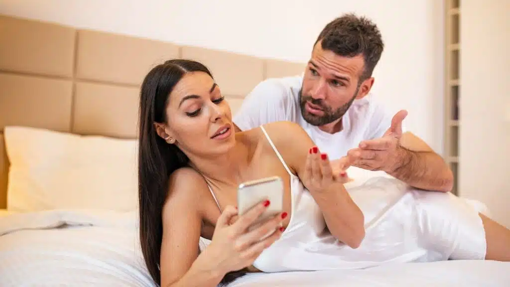 A woman lying on a bed looks at her phone while a man gestures toward her.