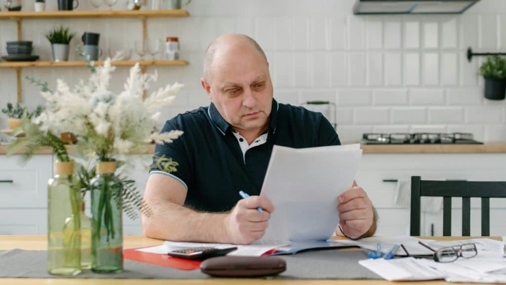 A man sits at a kitchen table holding a blue pen and reviewing several white documents.