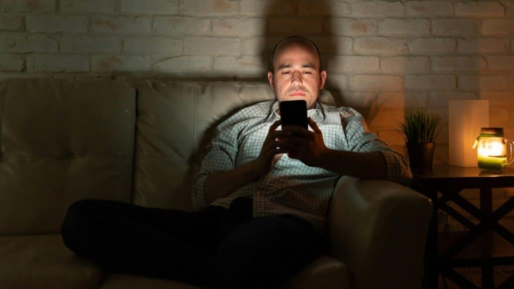 A man sits on a couch in a dark room looking at his illuminated smartphone screen.