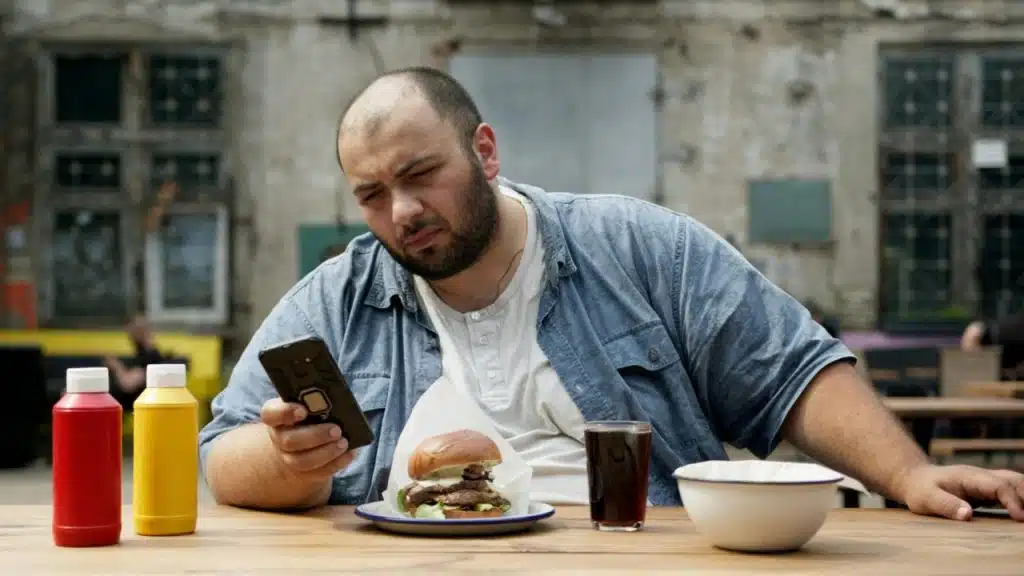 A man at an outdoor table looks at his phone while a burger sits before him.