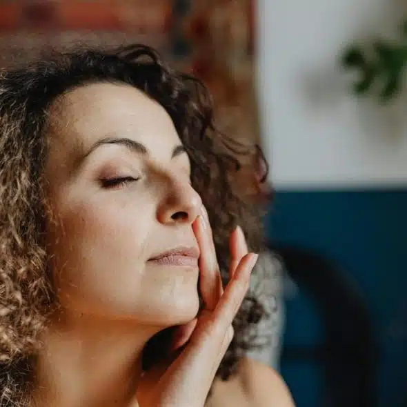 A woman with curly hair and closed eyes gently touching her face indoors.