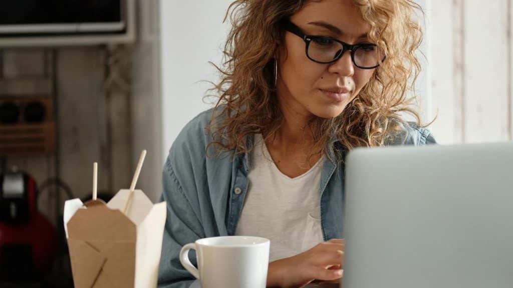 A woman with curly hair and glasses works on a laptop beside takeout food and a mug.