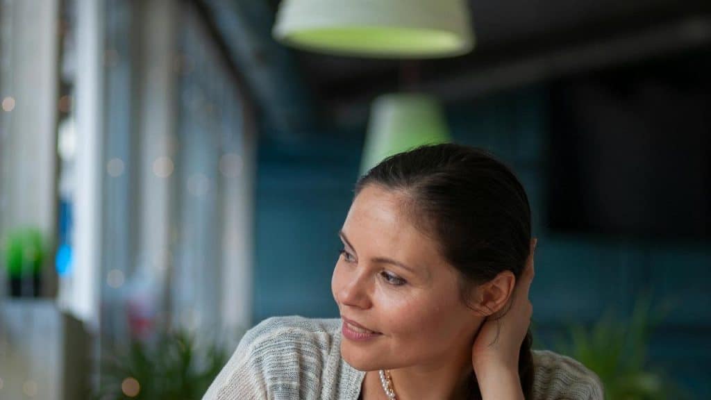 A woman sits indoors, smiling softly and touching her hair.
