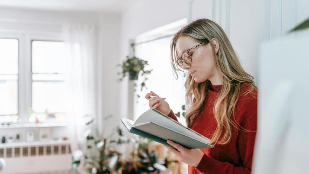 A woman wearing glasses reading a book and taking notes indoors.