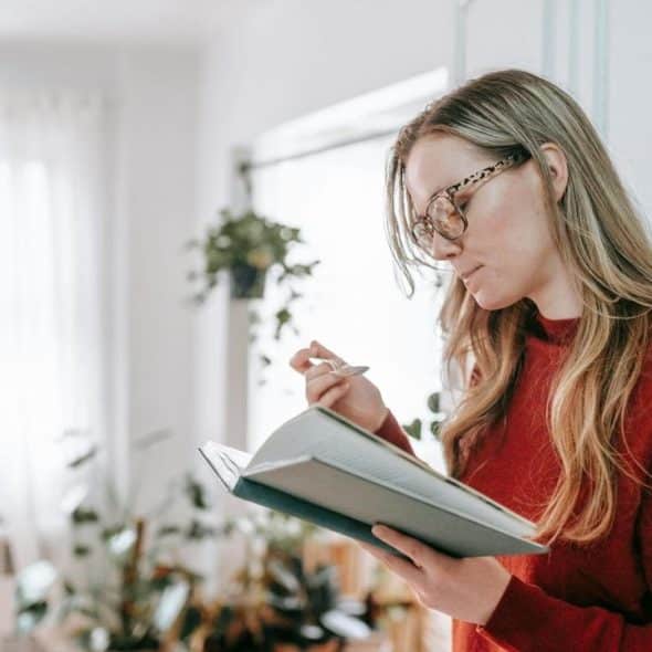 A woman wearing glasses reading a book and taking notes indoors.
