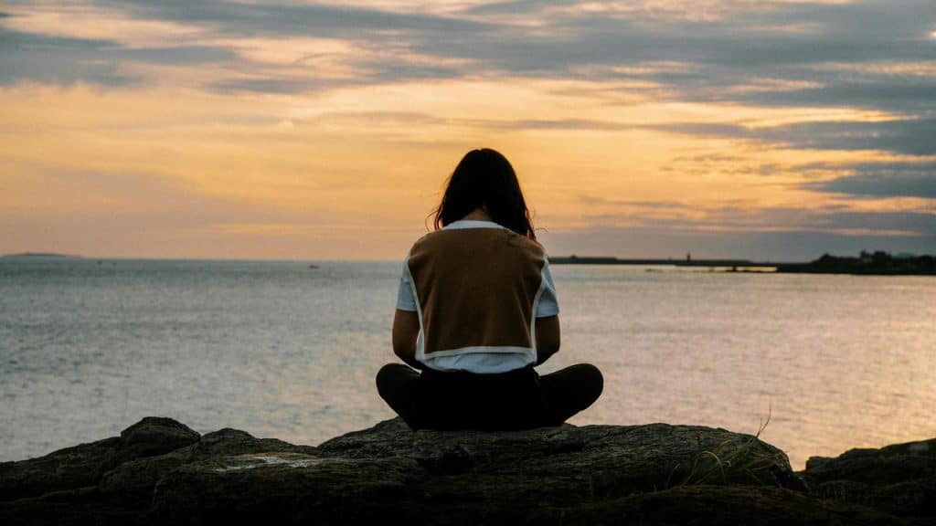 A person sitting cross-legged on a rock overlooking the ocean at sunset.