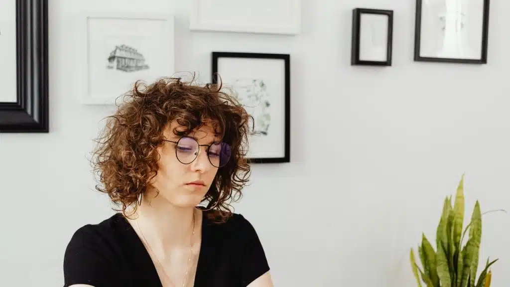 A person with curly hair and glasses sitting indoors near framed wall art and a plant.