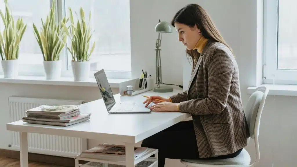 A woman working on a laptop at a desk with plants by a window.