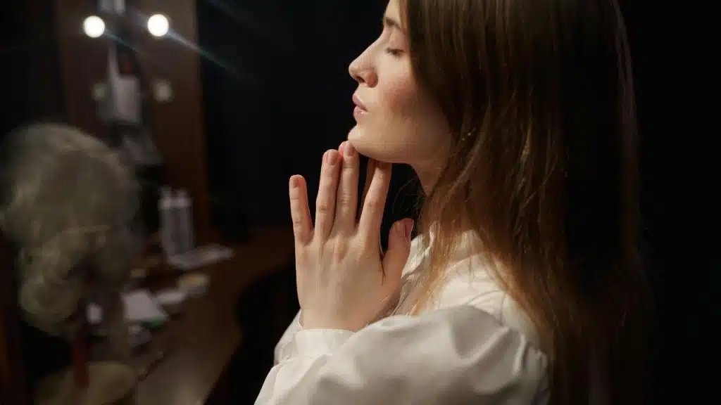A woman with hands clasped near her face standing in a dimly lit room.