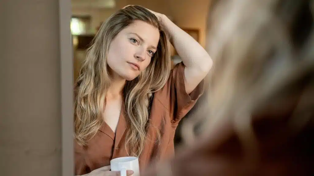 A woman holding a mug while adjusting her hair and looking in a mirror.