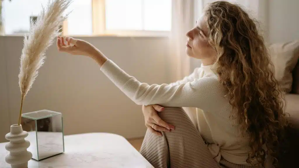 A woman sitting indoors gently touching a dried plant near a window.