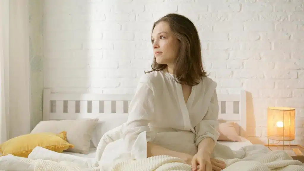 A woman sitting on a bed looking thoughtfully toward a window.