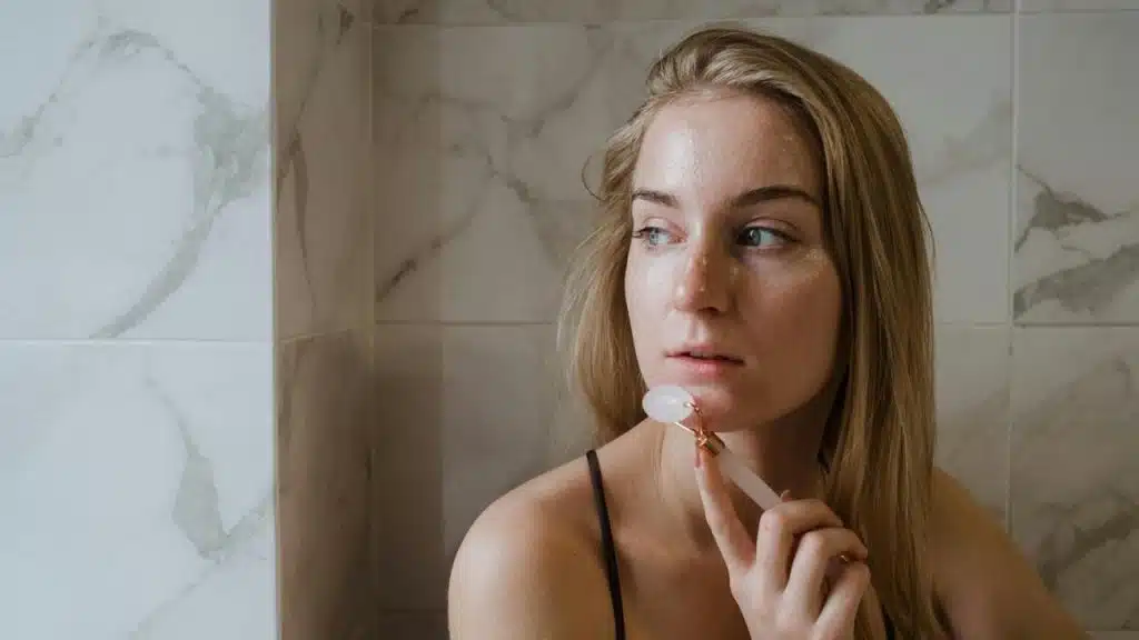 A woman using a facial roller on her chin while standing by a tiled wall.