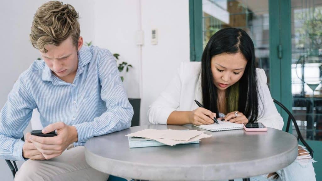 A man looking at his phone while a woman writes in a notebook at a table.
