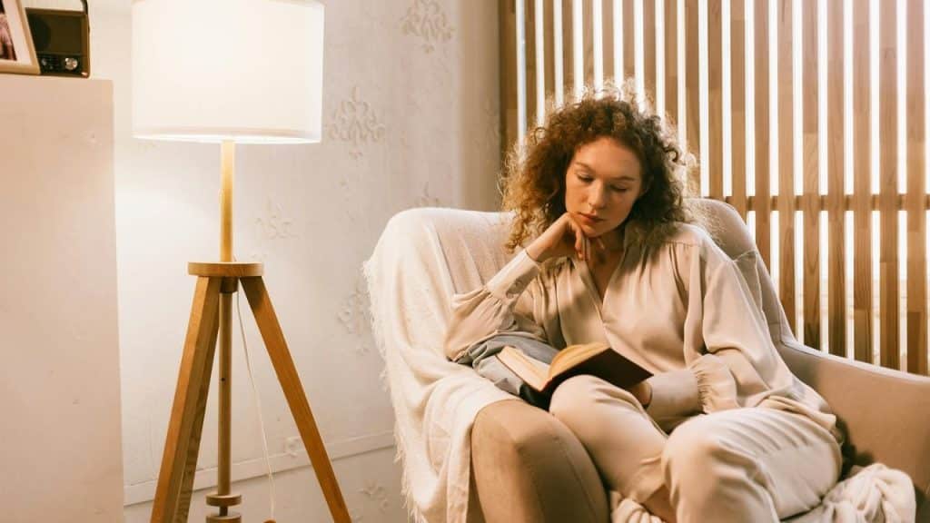 A woman sitting in an armchair reading a book beside a lamp.