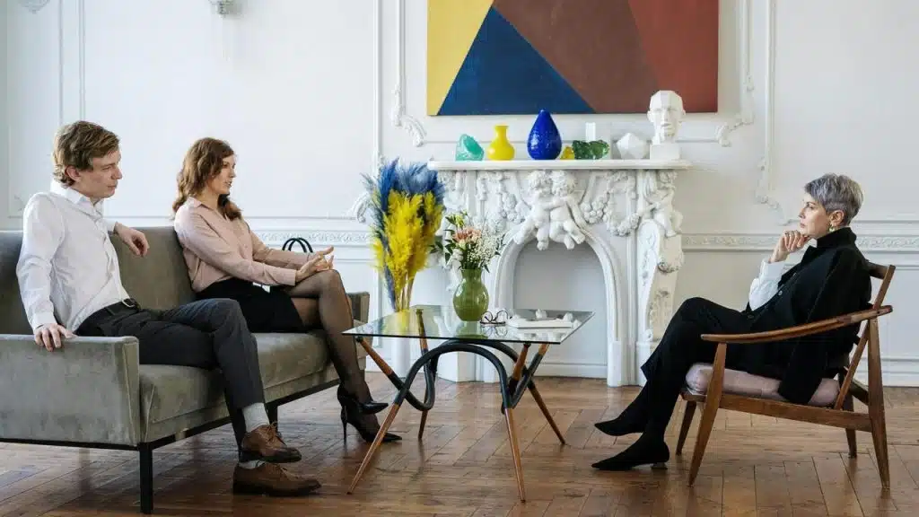 A couple sitting on a couch speaking with a therapist in a bright living room.