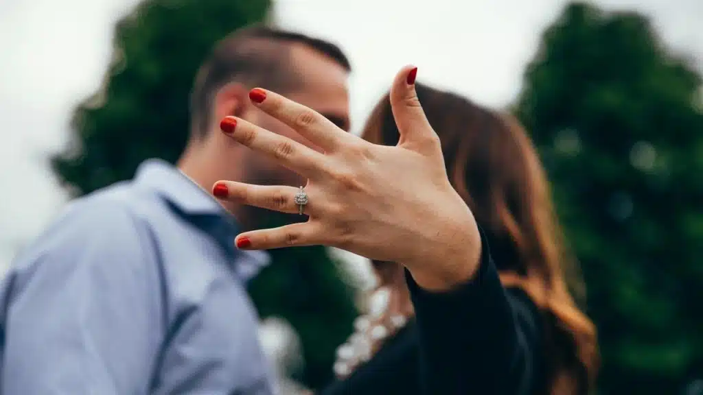 A hand showing an engagement ring with a blurred couple behind it.