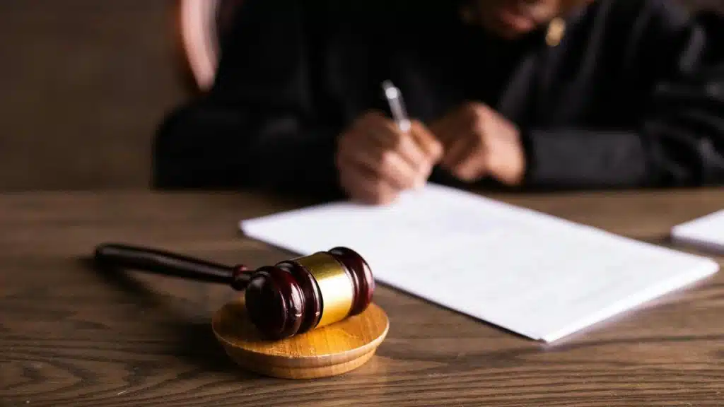 A judge’s gavel on a desk with a person signing papers in the background.