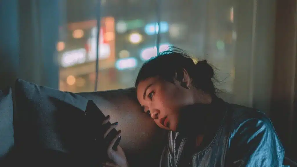 A woman sitting on a couch at night looking sadly at her phone.