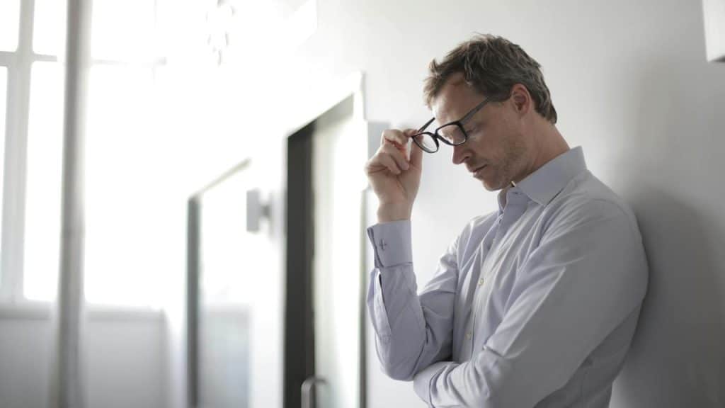 A worried man leaning against a wall while holding his glasses.