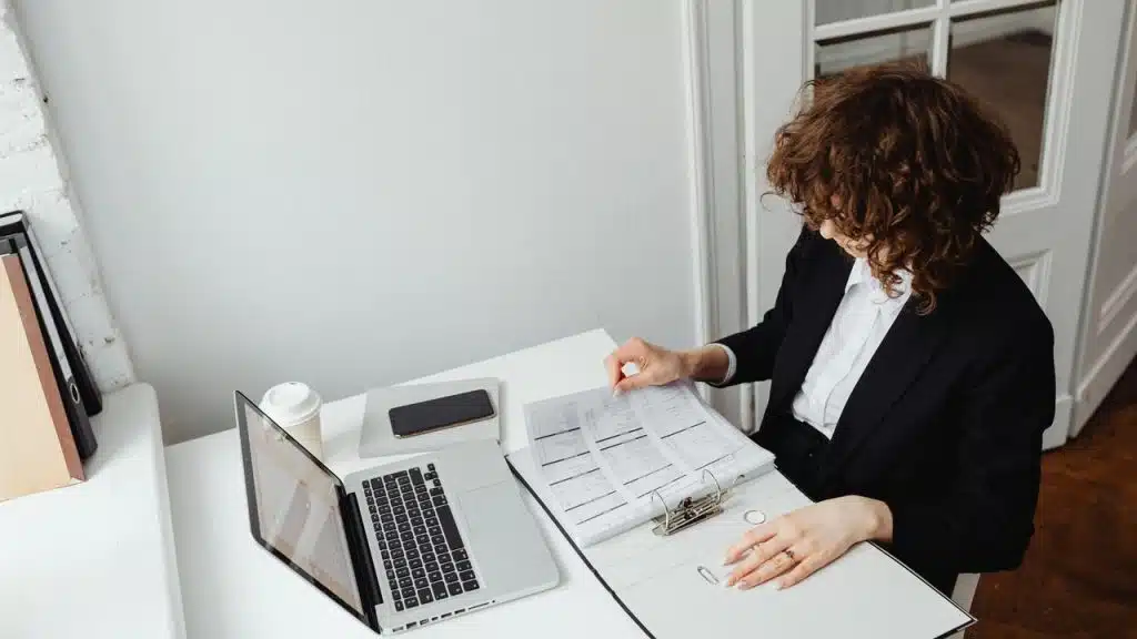 A woman reviewing documents at a desk with a laptop.