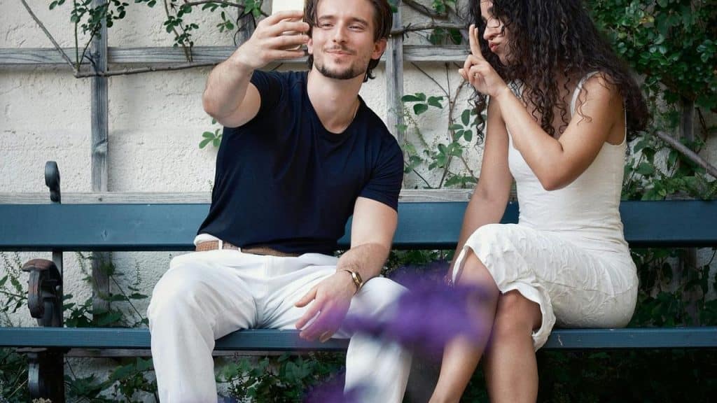 A man taking a selfie on a bench while a woman beside him makes a peace sign.