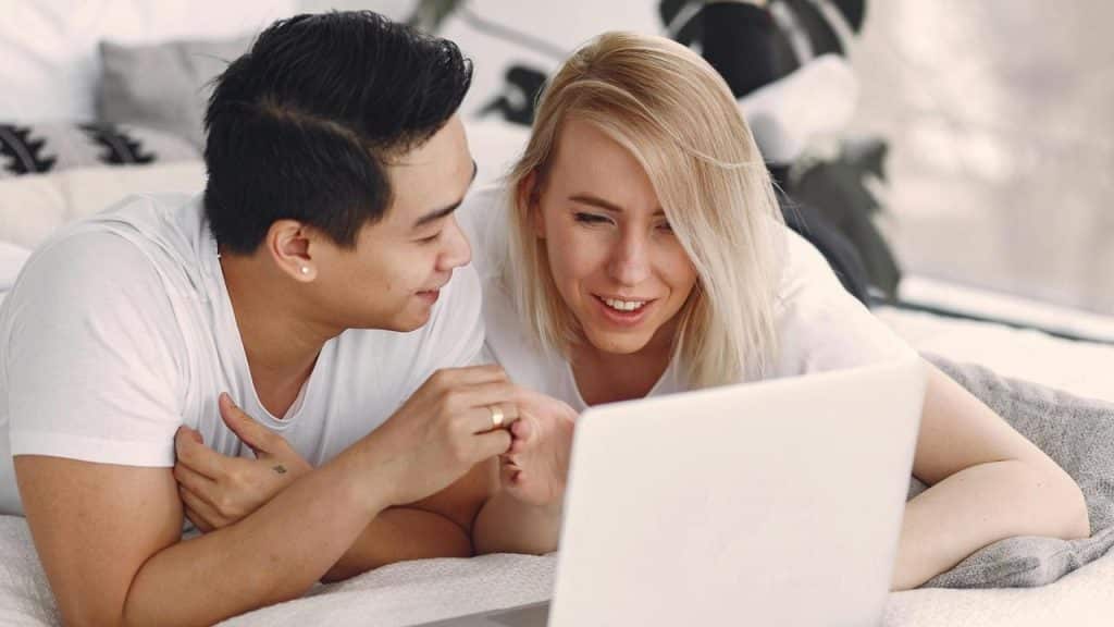 A couple lying on a bed and looking at a laptop together.
