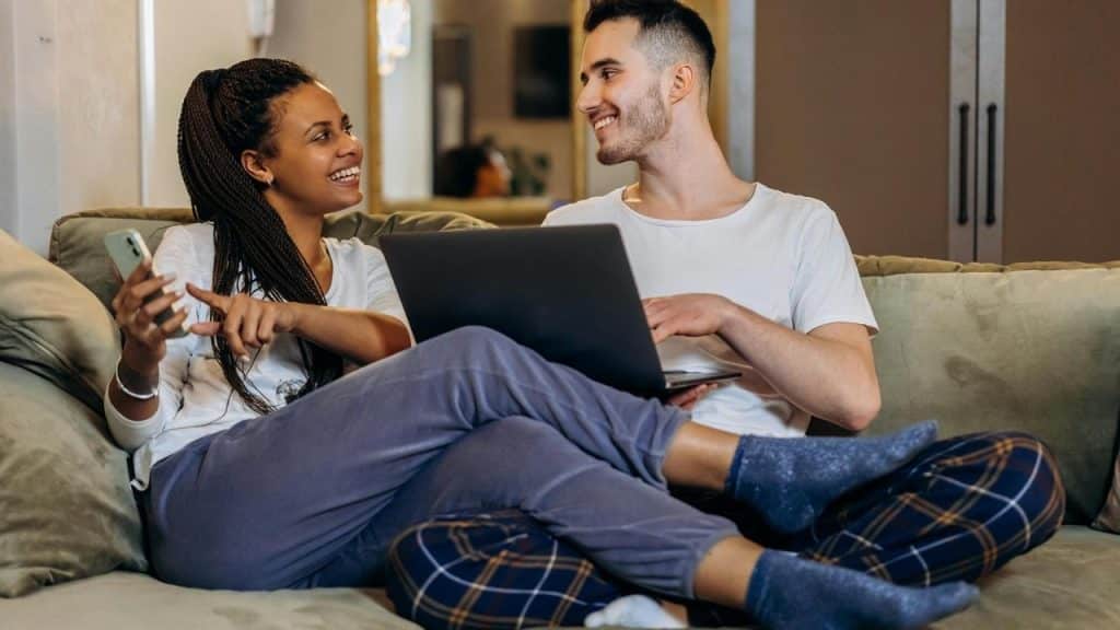 A couple sitting on a couch using a laptop and phone together.