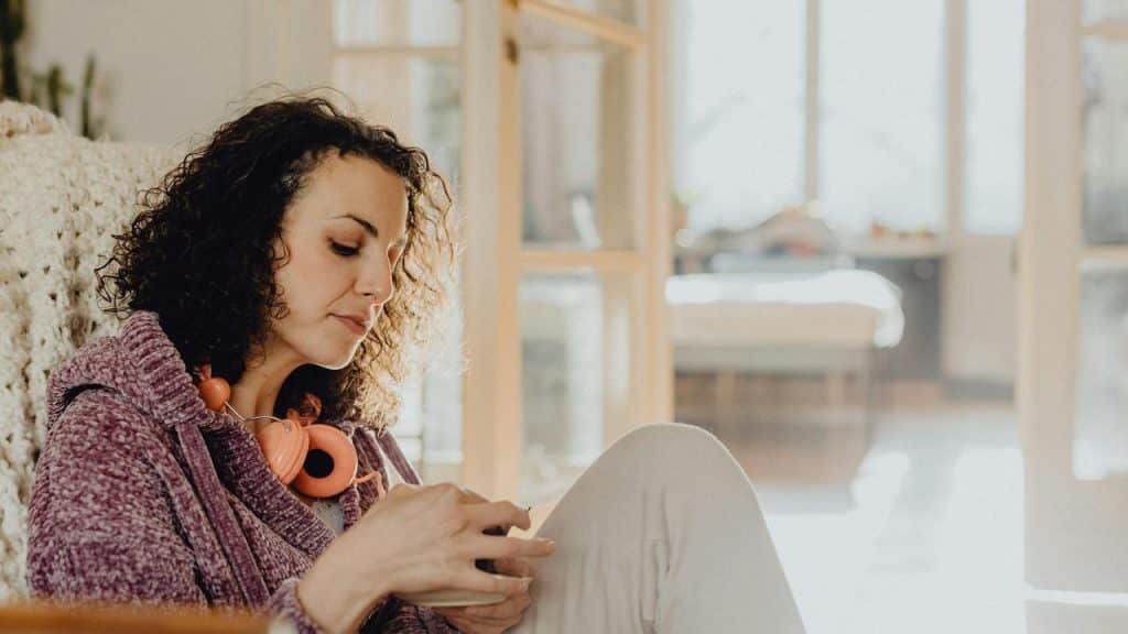 A woman sitting at home using her phone with headphones around her neck.