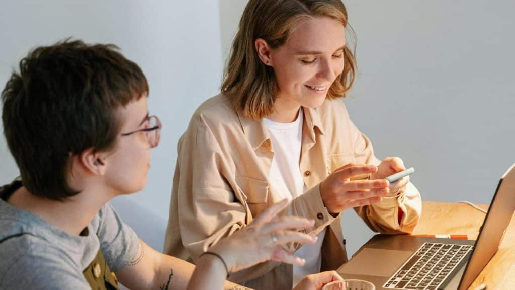 A smiling woman using a phone beside another person at a table with a laptop.