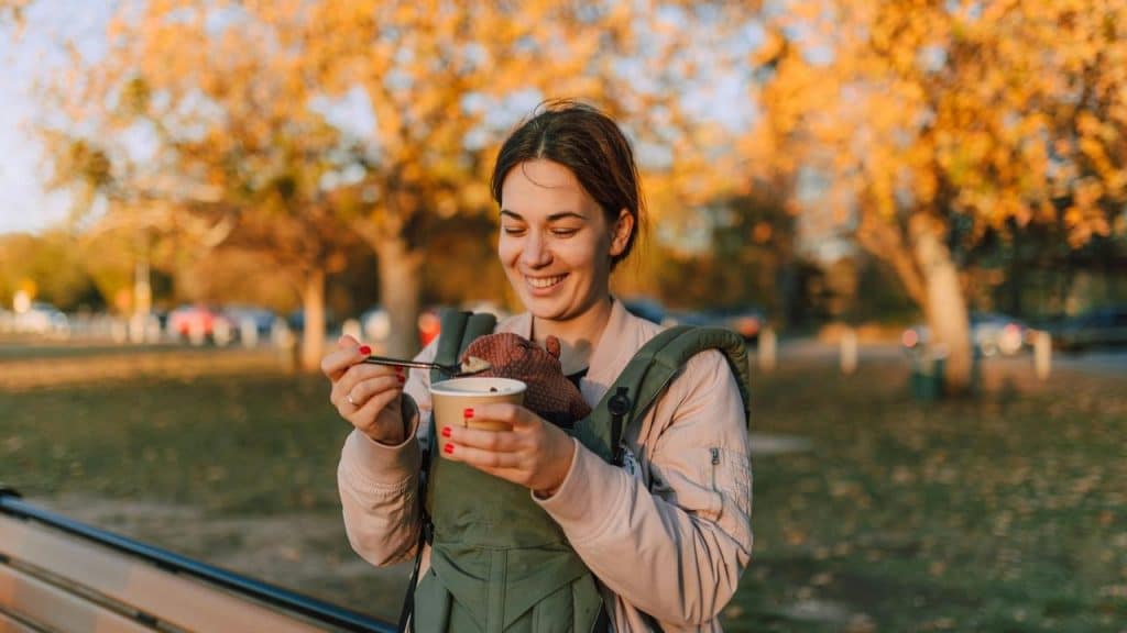 A woman feeding a baby in a carrier while standing in a park with autumn trees.