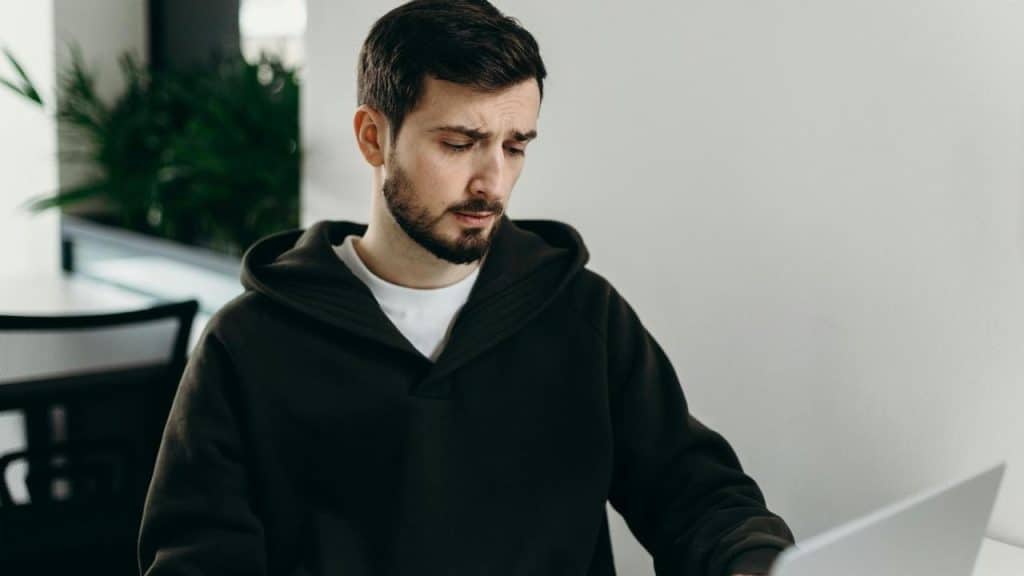A man in a hoodie looking concerned while using a laptop.
