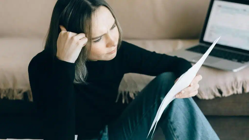 A woman looking concerned while reading a document with a laptop nearby.