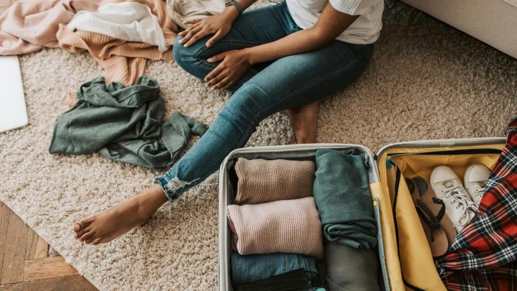 A person sitting on the floor packing clothes into an open suitcase.