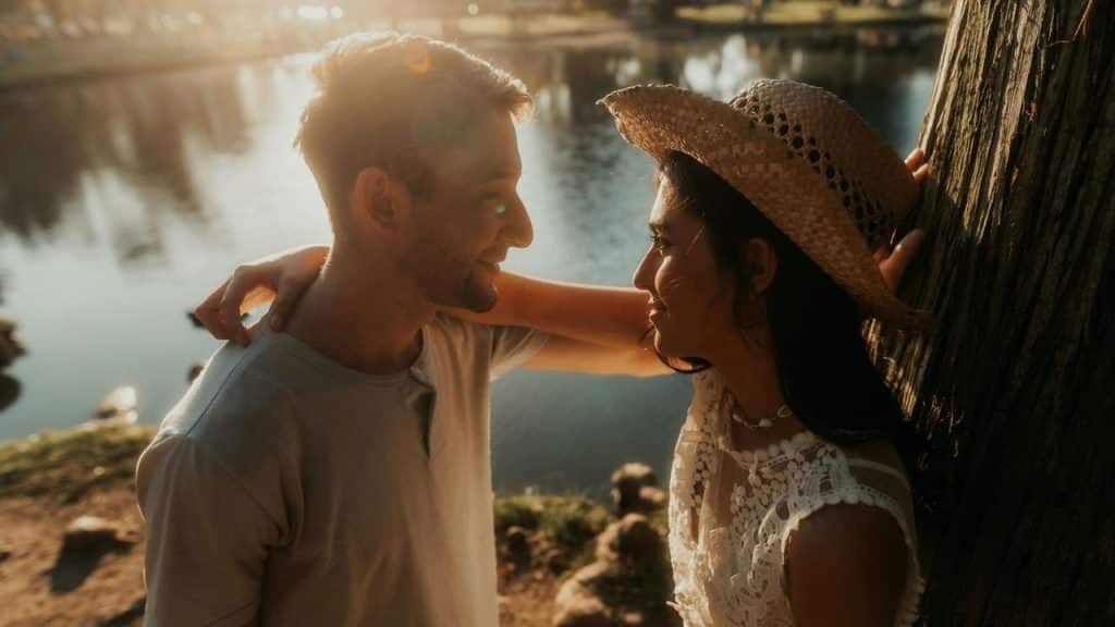 A couple standing by a lake at sunset, smiling at each other.