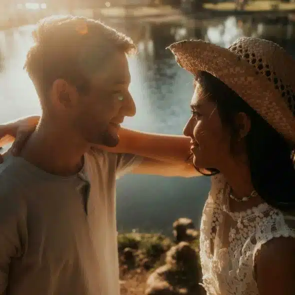 A couple standing by a lake at sunset, smiling at each other.