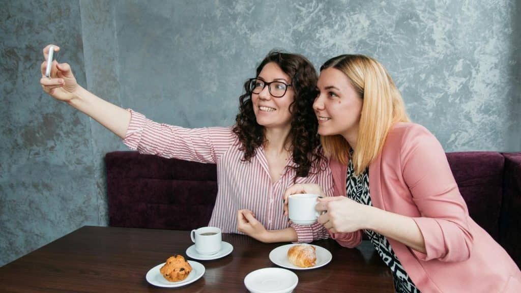 A pair of women sitting at a café table with coffee and pastries, taking a selfie together.