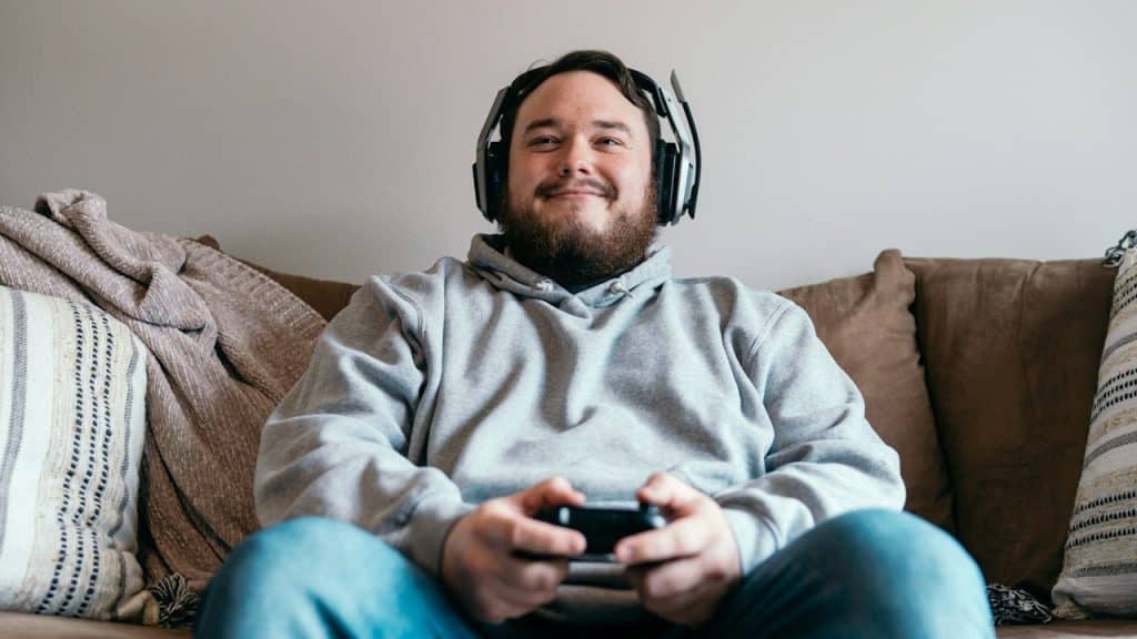 A bearded man smiling while playing a video game and wearing headphones as he sits on a couch.