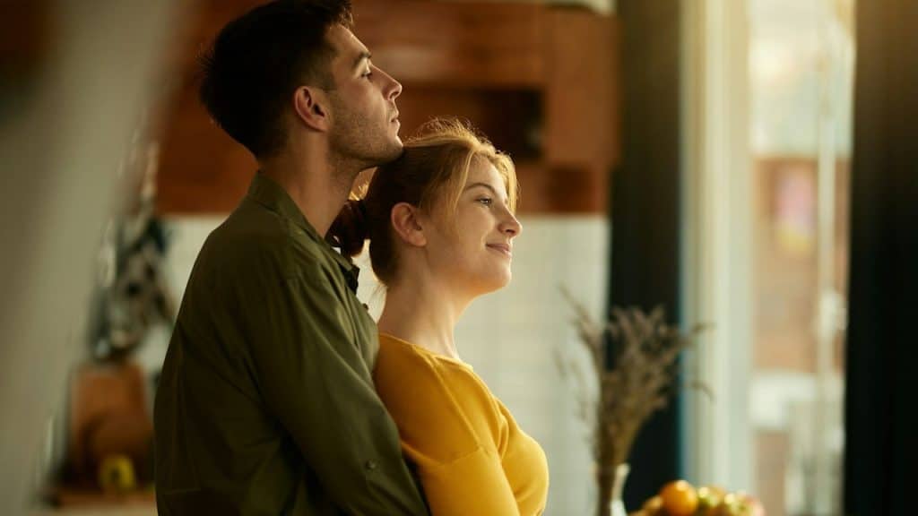 Smiling woman daydreaming while her boyfriend is standing behind and embracing her at home.