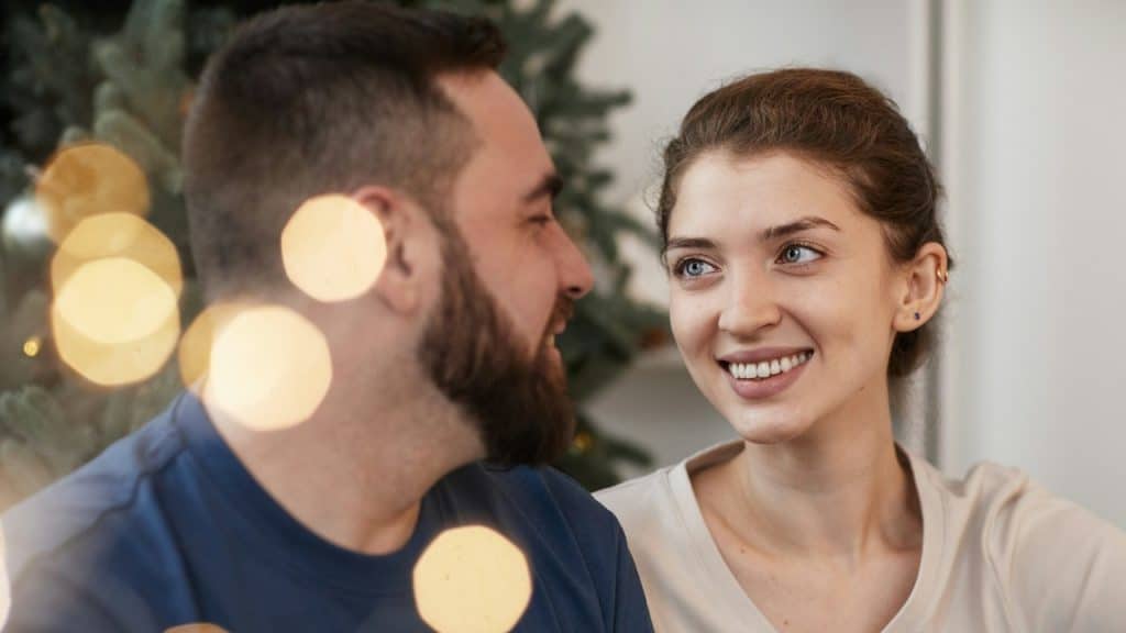 Happy young Caucasian woman looking with love at boyfriend while they are celebrating Christmas together with lens flare.