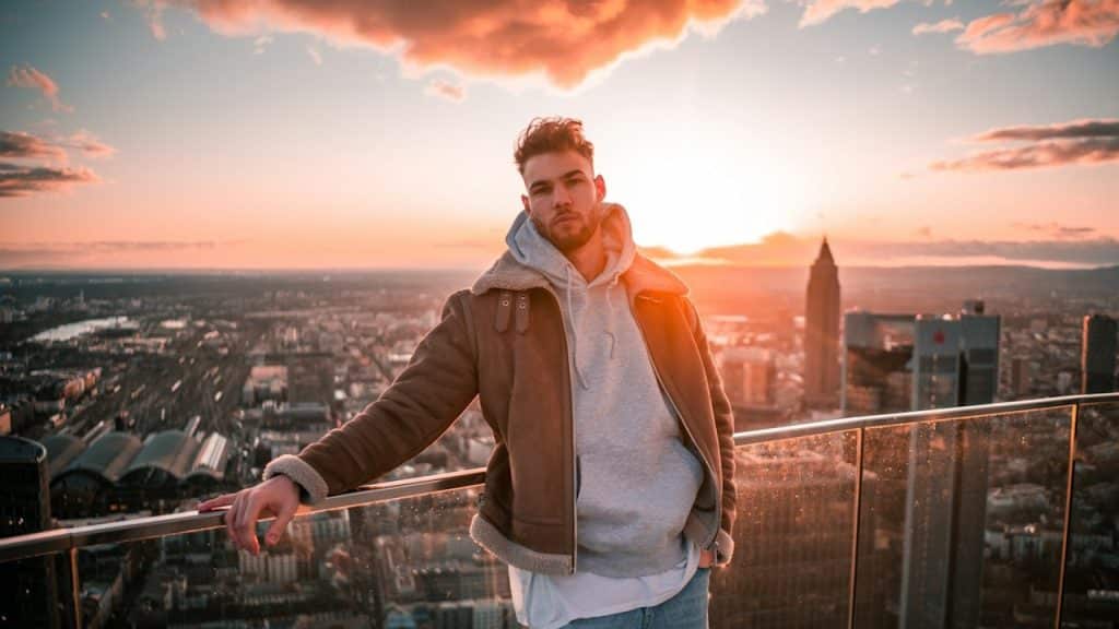 A bearded man standing on a balcony overlooking an urban area.