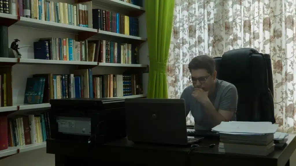 A man with glasses sits at a desk, looking intently at his laptop.
