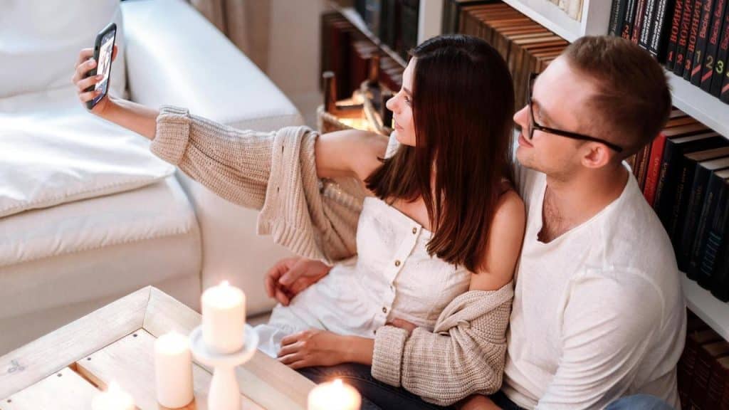A couple sitting on the floor by a couch and bookshelf, taking a selfie together.