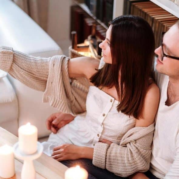 A couple sitting on the floor by a couch and bookshelf, taking a selfie together.
