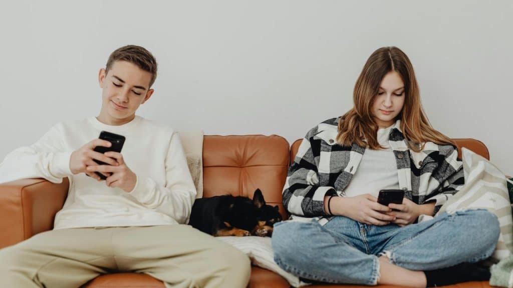 A couple sitting on a couch using their phones with a dog between them.